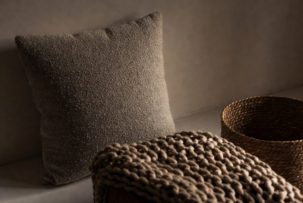 Neutral room layered with boucle cushion, chunky knit throw and woven basket showing rich textures, warm light
