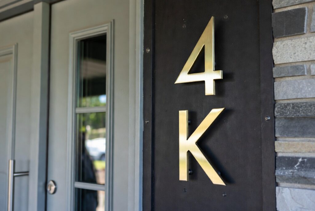 : Modern home entry with oversized brushed brass house numbers on dark wall next to clean front door, natural daylight