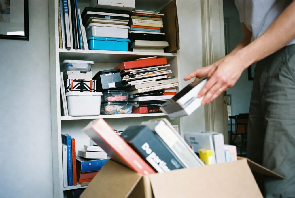 Person decluttering overstuffed shelf by placing items into donation box, natural home lighting, candid organizing scene