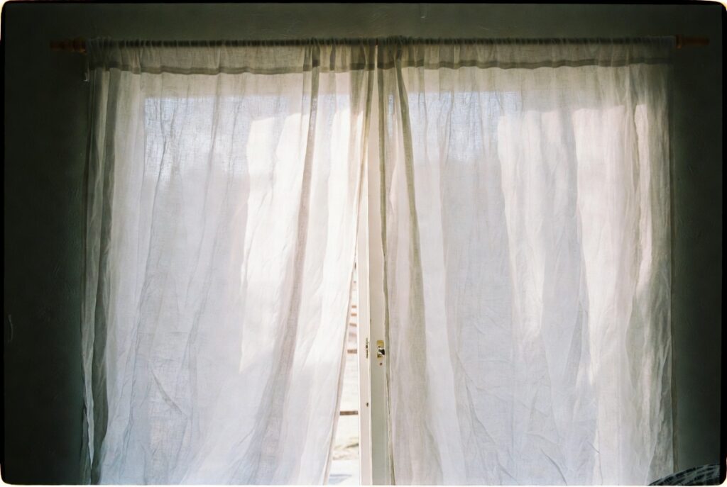 Sheer white linen curtains billowing gently at open window with soft daylight filling the room, airy natural light
