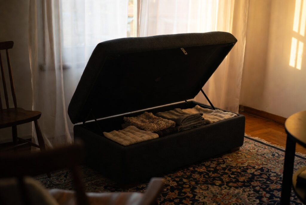 Living room ottoman with lift-top storage revealing neatly folded blankets inside, warm afternoon light, organized home