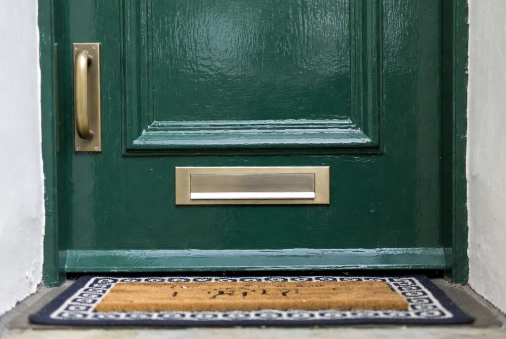 Close-up home entry with brushed brass door handle, knocker and letterbox on bold green door, woven doormat, natural light