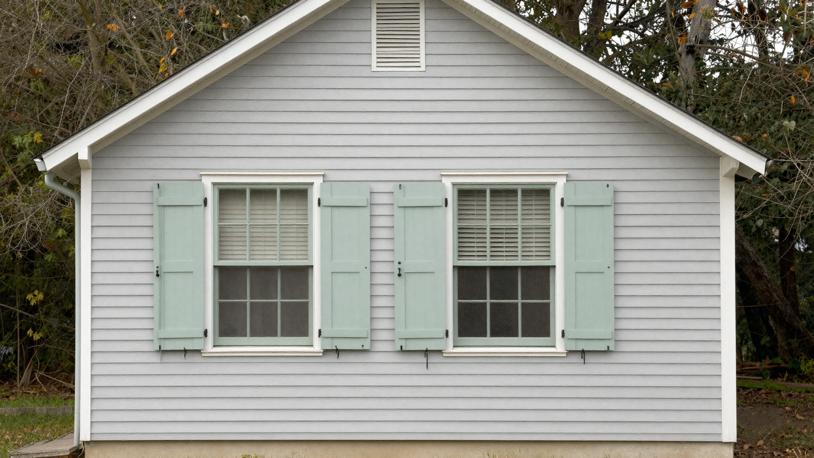 House exterior with light gray siding and soft sage green shutters blending with tree-wrapped surroundings